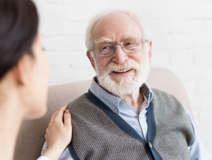 A smiling elderly man with white hair and glasses looks warmly at a younger caregiver who gently places a hand on his shoulder, illustrating compassionate memory care support.
