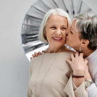 Two older women sharing a joyful moment together — one smiling brightly as the other kisses her cheek — representing LGBTQ+ connection and belonging in senior living communities.