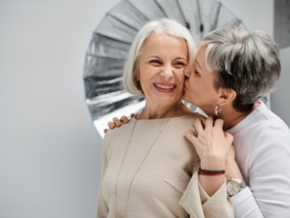 Two older women sharing a joyful moment together — one smiling brightly as the other kisses her cheek — representing LGBTQ+ connection and belonging in senior living communities.
