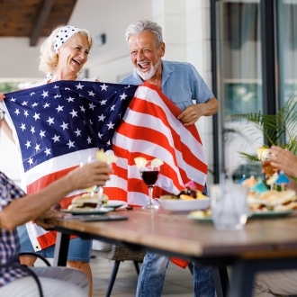 Seniors at a senior living community laughing together while holding an American flag at a Fourth of July gathering, celebrating independence and connection.