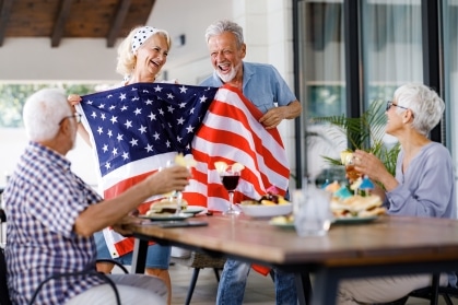 Seniors at a senior living community laughing together while holding an American flag at a Fourth of July gathering, celebrating independence and connection.