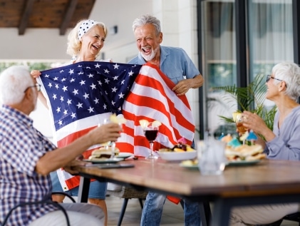 Seniors at a senior living community laughing together while holding an American flag at a Fourth of July gathering, celebrating independence and connection.