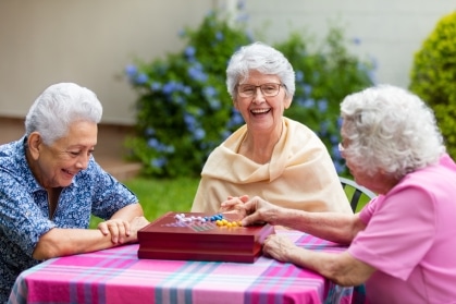 Three smiling senior women play a wooden marble board game together at an outdoor table on a sunny afternoon