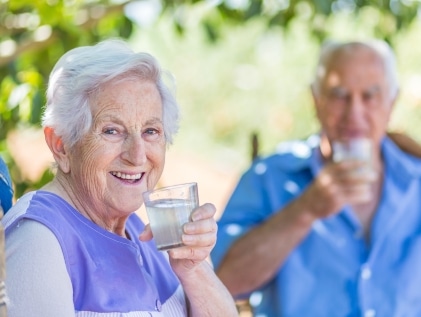 Smiling senior woman drinks a glass of water outdoors in the shade while an older man hydrates in the background
