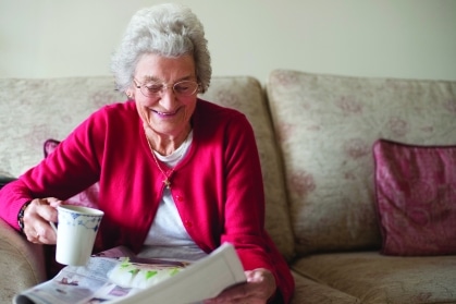 Smiling senior woman in a red cardigan enjoys her morning coffee and newspaper, a familiar part of her daily routine.