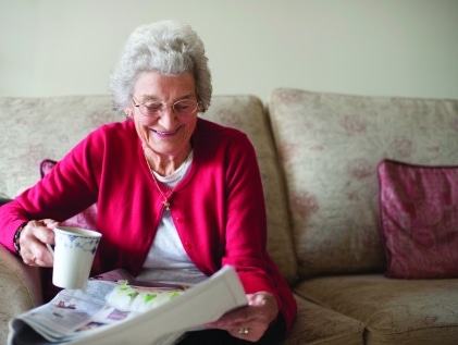 Smiling senior woman in a red cardigan enjoys her morning coffee and newspaper, a familiar part of her daily routine.