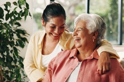 Adult daughter with her arm around her smiling senior mother as they laugh together in a bright memory care community living room