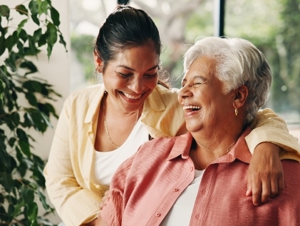 Adult daughter with her arm around her smiling senior mother as they laugh together in a bright memory care community living room