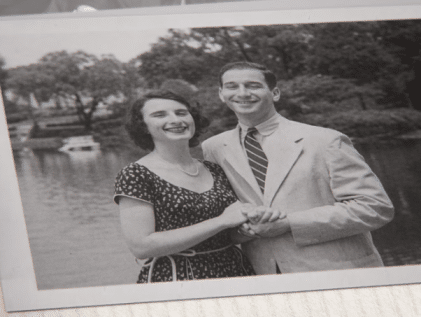 Vintage black-and-white photo of a smiling young couple holding hands beside a tree-lined pond, the kind of old family photo AI tools can restore to spark reminiscence.