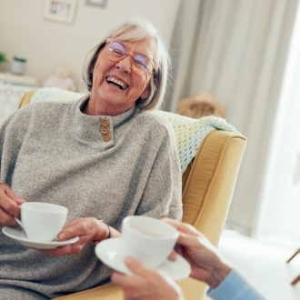 Two senior women laugh together over cups of tea in a bright, comfortably furnished assisted living apartment