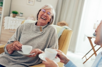 Two senior women laugh together over cups of tea in a bright, comfortably furnished assisted living apartment