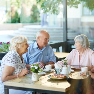 Four older adults chatting and laughing over tea and pastries at an outdoor café table, a social outing that supports brain health in older adults.