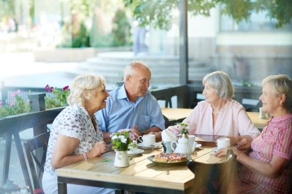 Four older adults chatting and laughing over tea and pastries at an outdoor café table, a social outing that supports brain health in older adults.