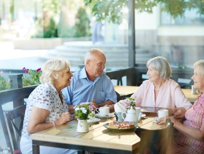 Four older adults chatting and laughing over tea and pastries at an outdoor café table, a social outing that supports brain health in older adults.