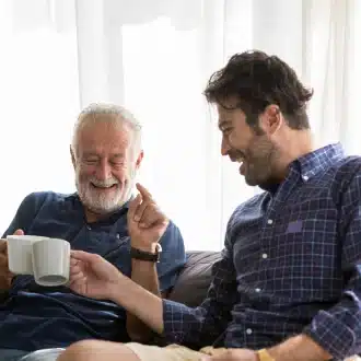 Smiling senior father and adult son laughing together on a couch, clinking coffee mugs in a bright living room.