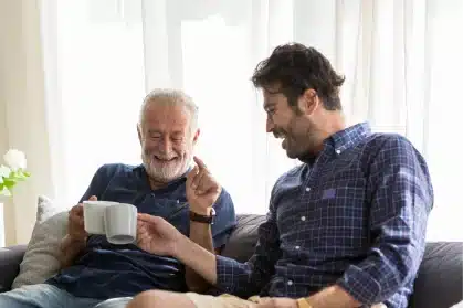 Smiling senior father and adult son laughing together on a couch, clinking coffee mugs in a bright living room.