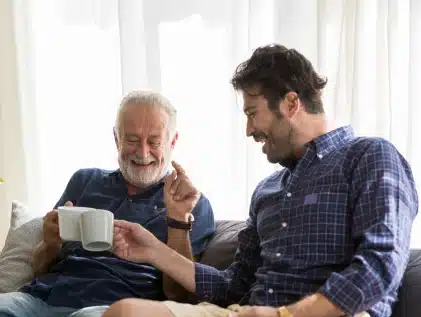 Smiling senior father and adult son laughing together on a couch, clinking coffee mugs in a bright living room.