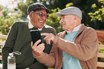 Two smiling older men wearing flat caps sit on a garden bench looking at a smartphone together, enjoying the easy friendship that grows in a senior living community.