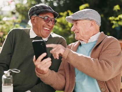 Two smiling older men wearing flat caps sit on a garden bench looking at a smartphone together, enjoying the easy friendship that grows in a senior living community.